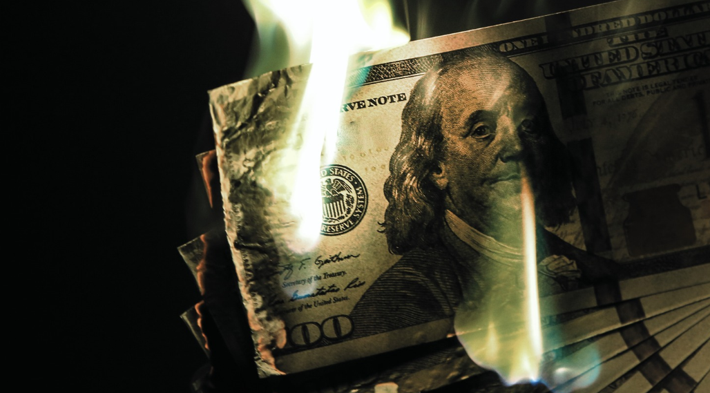 Stack of hundred-dollar bills partially burning with flames against a dark background.