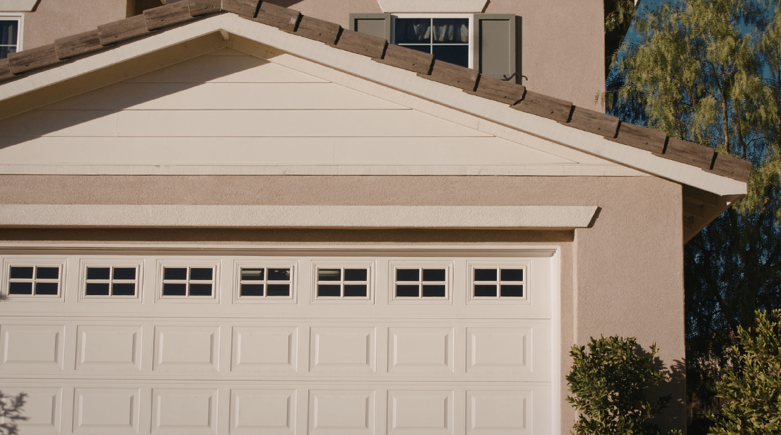 A beige house with a white garage door featuring small rectangular windows, a gabled roof with brown shingles, an upstairs window with green shutters, and bushes near the entrance—ideal for proactive termite control to prevent termite infestation.